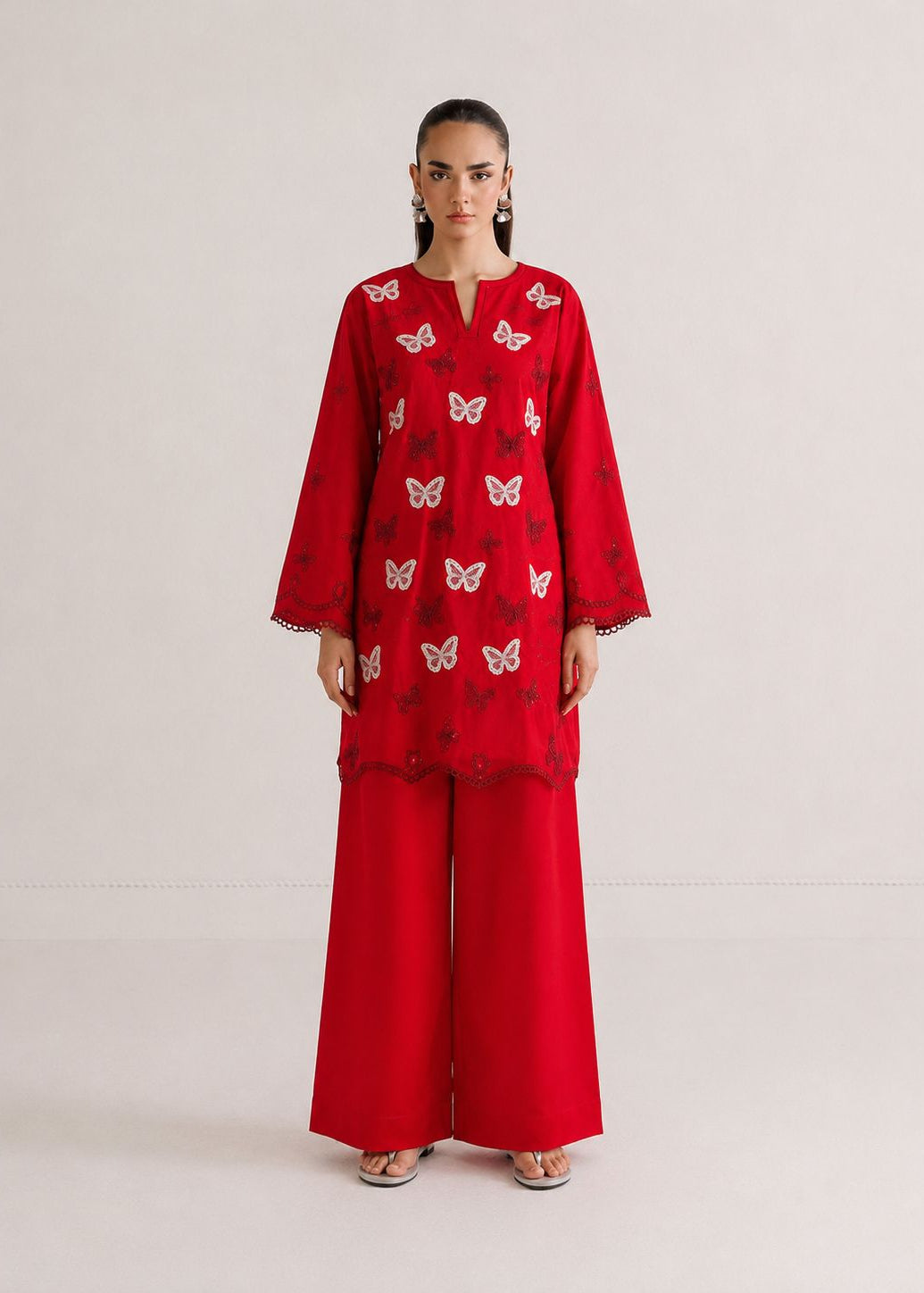 A model stands against a plain light beige studio background wearing a bright red traditional outfit consisting of a long-sleeve embroidered kurta with butterfly motifs and matching wide-leg trousers.