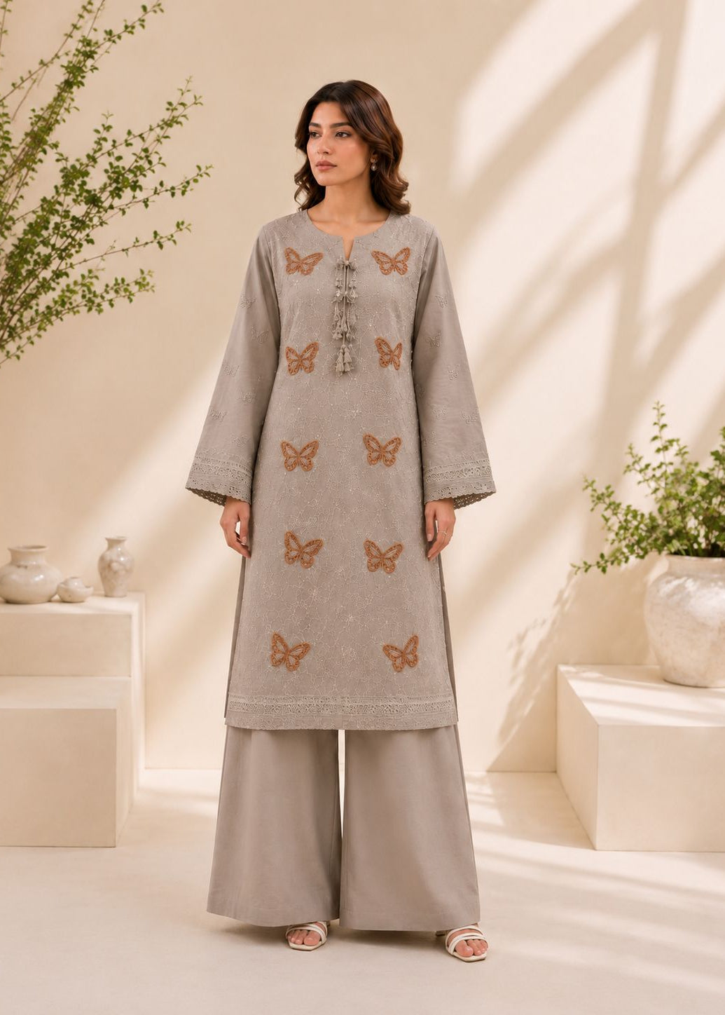 A woman models a light gray three-piece kurta set with matching wide-leg trousers. The kurta features embroidered brown butterflies and a tassel tie at the neckline.