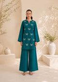 A woman stands in a warm beige studio setting wearing a teal two-piece outfit with embroidered butterfly motifs on the kurta and matching wide-leg pants.