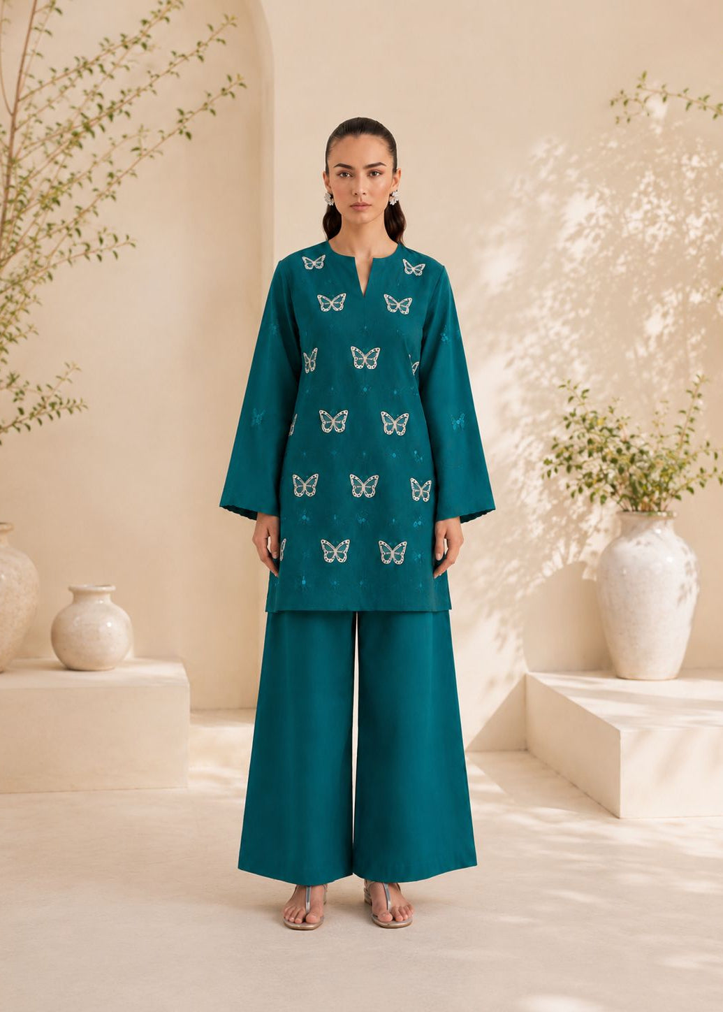 A woman stands in a warm beige studio setting wearing a teal two-piece outfit with embroidered butterfly motifs on the kurta and matching wide-leg pants.