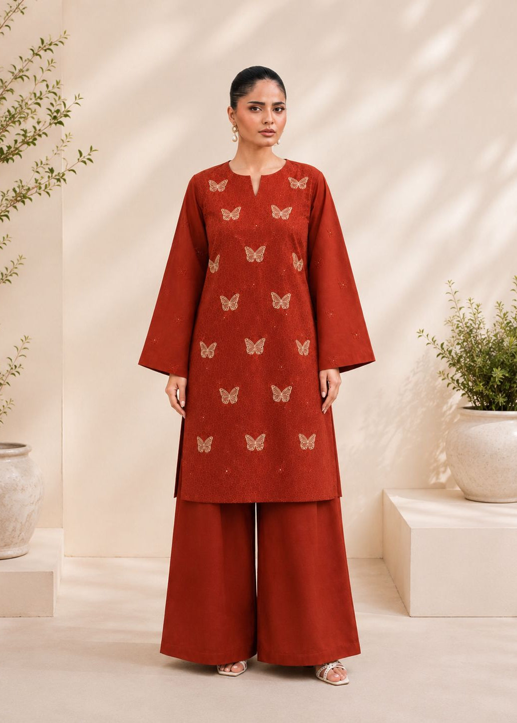 A woman stands in a studio setting wearing a rust-red kurta with small butterfly motifs and matching wide-leg pants. The background is a warm beige with decorative plants and soft shadows.