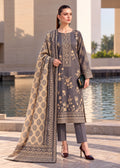 A woman models a three-piece traditional South Asian outfit consisting of a dark gray kurta with beige floral embroidery, matching gray trousers, and a beige dupatta with a gray geometric border, set against a modern outdoor background with a pool of water.