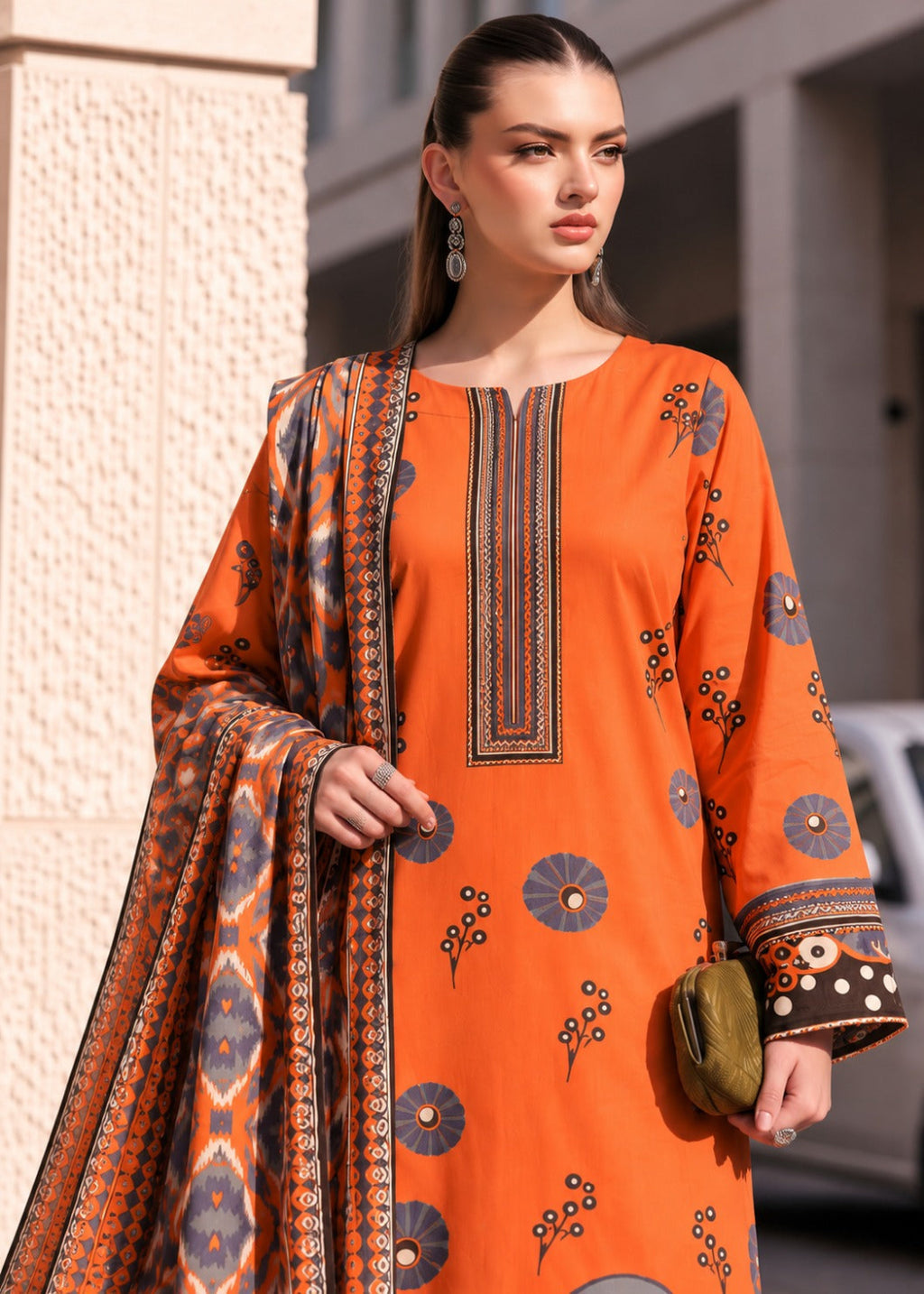 A woman modeling a bright orange traditional outfit with embroidered details and a patterned dupatta, standing outdoors against a light-colored building.