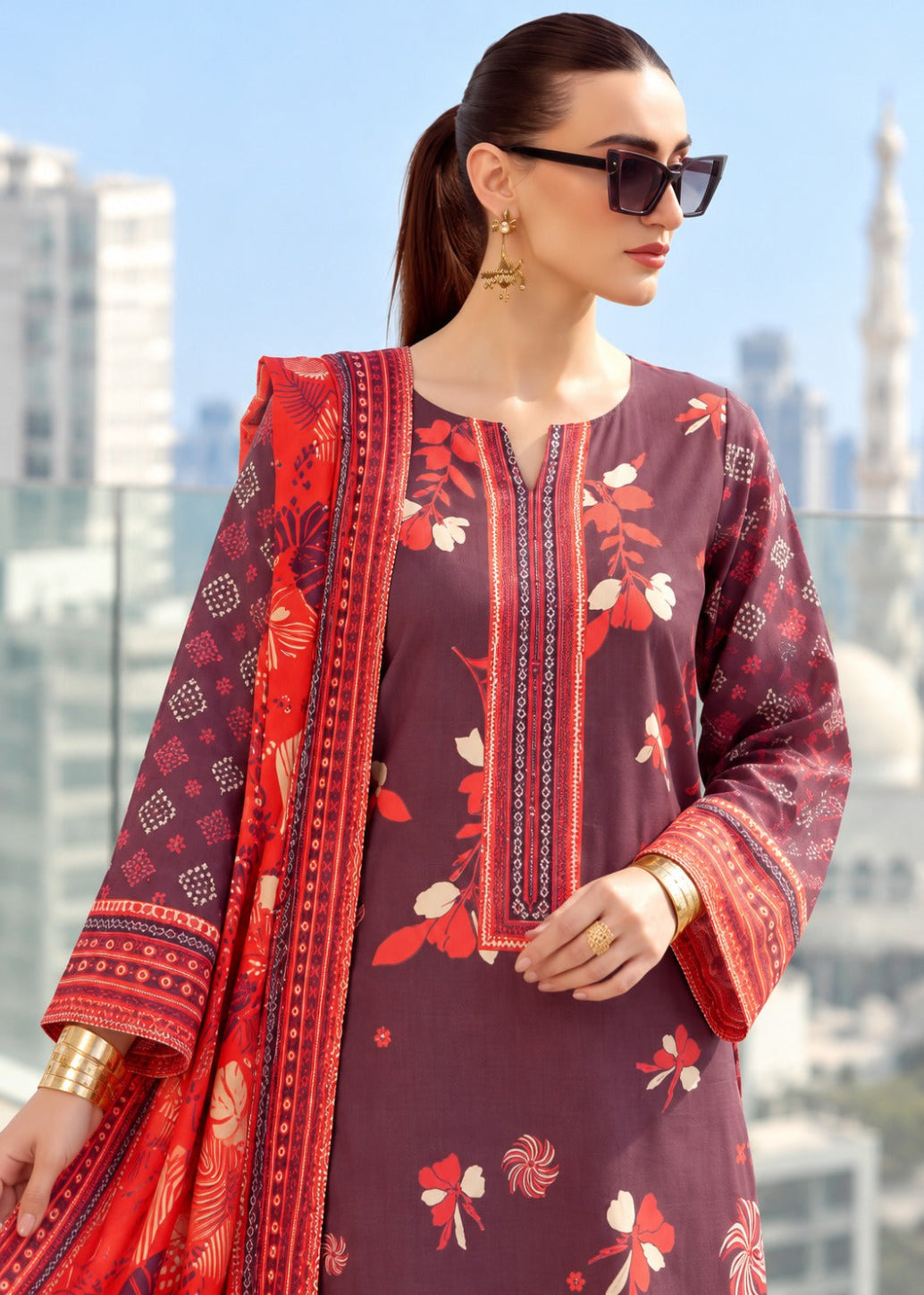 A woman wearing a traditional three-piece salwar kameez suit in deep plum purple, featuring red and cream floral prints, a matching red dupatta, and gold jewelry, set against a blurred city background.