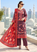 A woman models a three-piece salwar kameez suit set on a balcony with a city skyline and mosque in the background. The set features a deep plum purple kameez kurta with red and white floral embroidery, matching wide-leg trousers, and a vibrant red dupatta with gold and navy patterns.