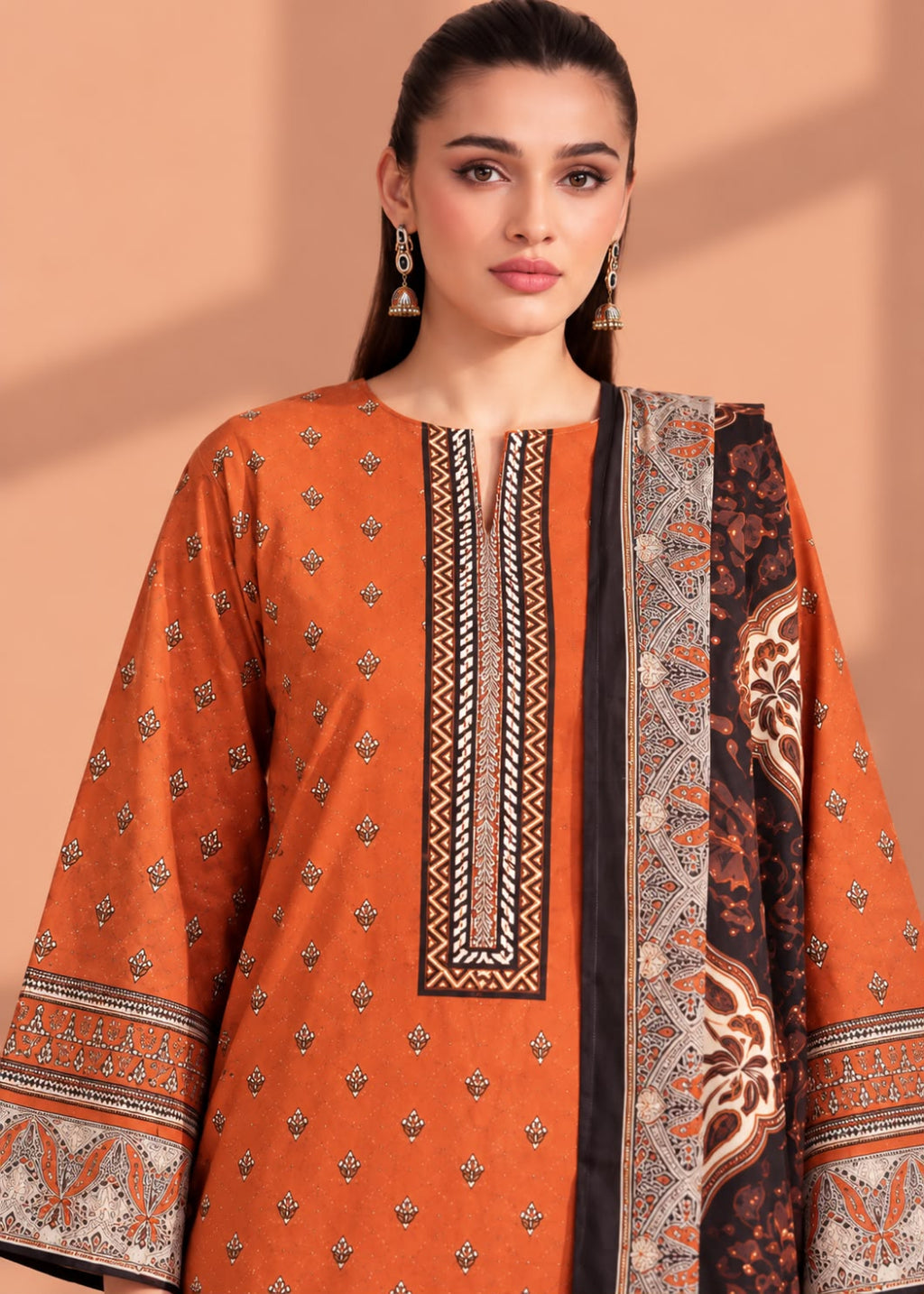 A woman modeling a traditional South Asian salwar kameez set in burnt orange, featuring a printed border dupatta and detailed geometric embroidery on the neckline.
