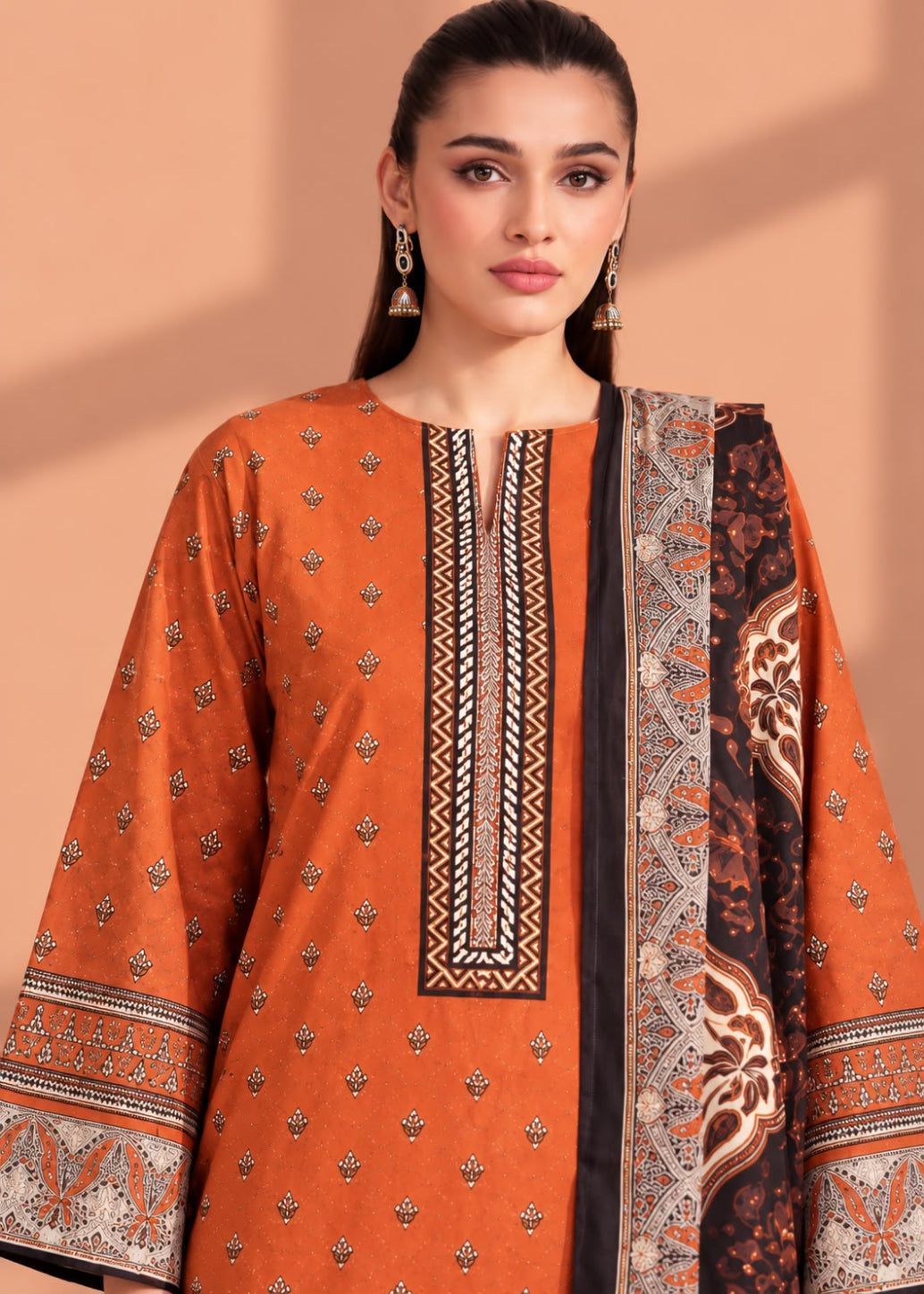 A woman modeling a traditional South Asian salwar kameez set in burnt orange, featuring a printed border dupatta and detailed geometric embroidery on the neckline.