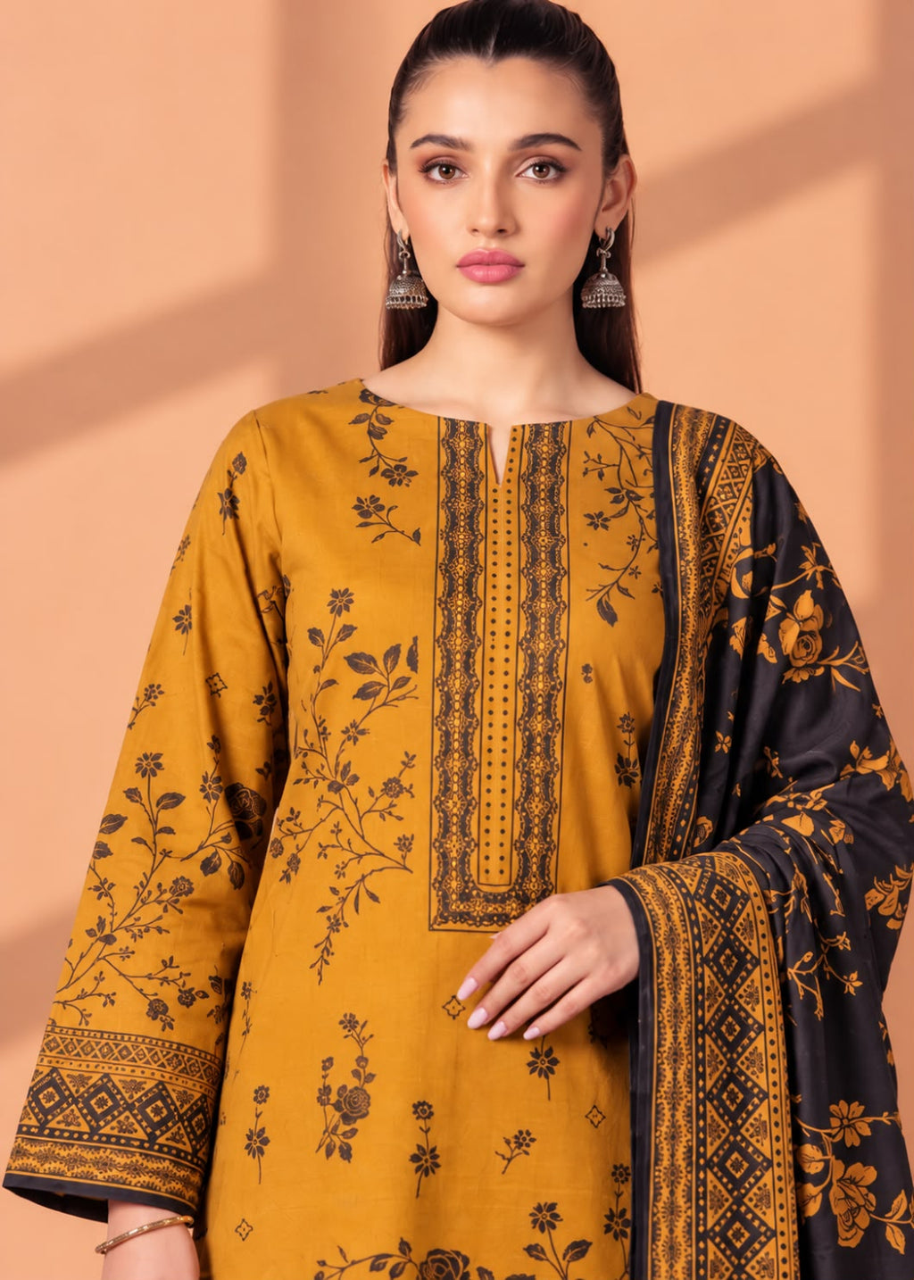 A woman modeling a mustard yellow ethnic kurta set with black floral prints and a black dupatta featuring gold floral and geometric borders, set against a peach-colored background with soft shadows.