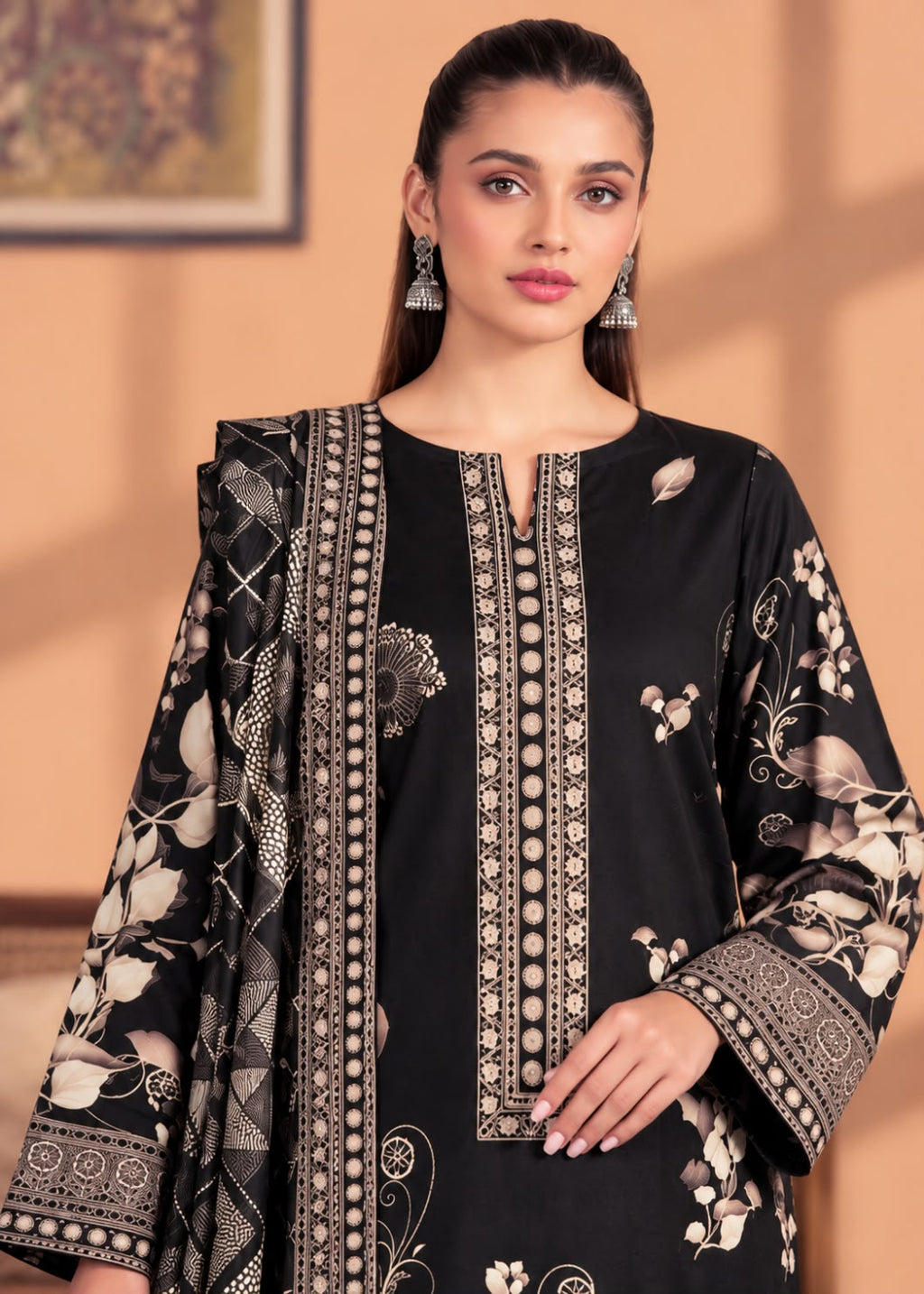 A woman modeling a black traditional outfit with beige and white floral embroidery and ornate border patterns, paired with a matching sheer dupatta, against a warm peach-colored background.