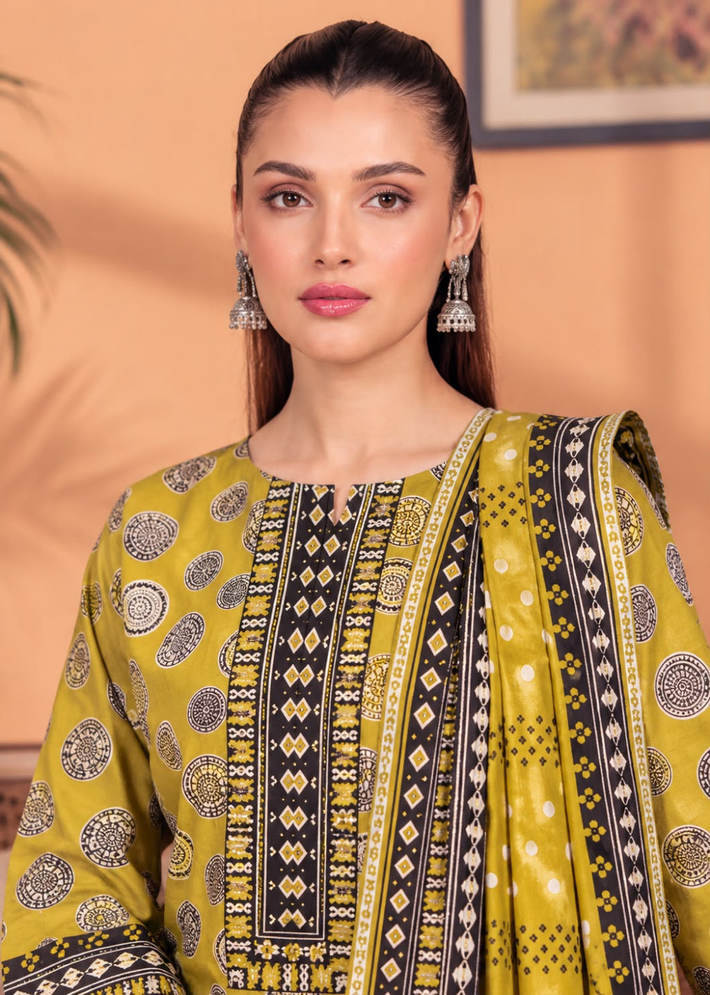 A woman wearing a yellow and black printed traditional outfit with a matching dupatta, featuring geometric and circular patterns, set against a warm peach-colored background.