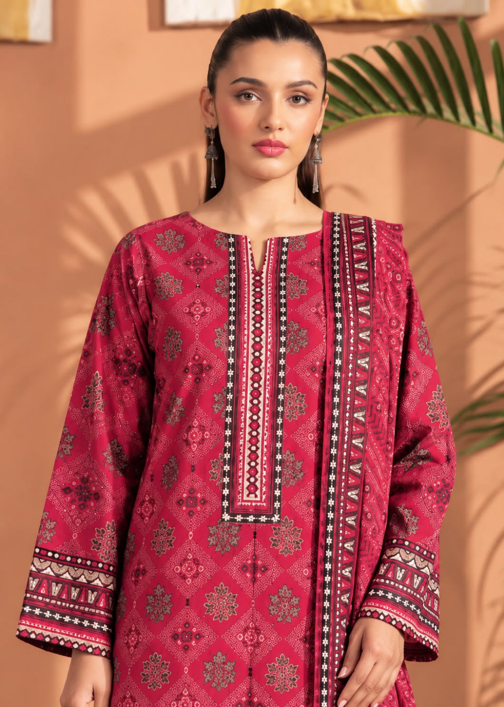 A woman modeling a traditional Indian salwar kameez set in a warm studio setting. The outfit is a deep magenta pink with intricate gold and black geometric patterns and border embroidery.