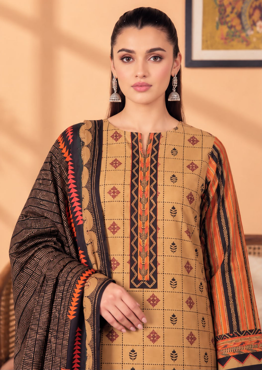 A woman modeling a traditional South Asian salwar kameez set featuring a beige kurta with geometric patterns, orange and black striped sleeves, and a dark brown dupatta with orange and black borders.