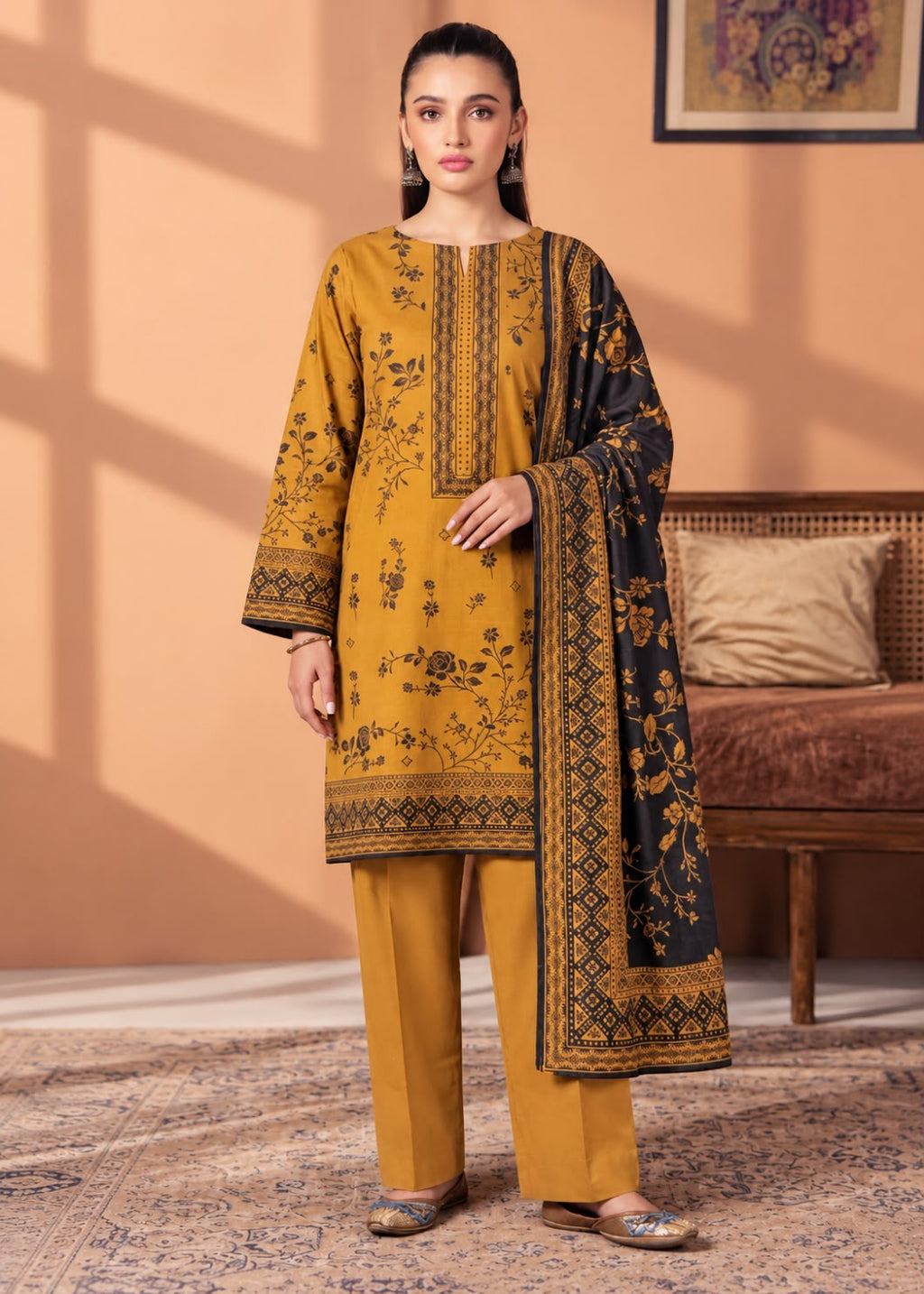 A woman models a traditional three-piece salwar kameez suit in mustard yellow and black. The kurta features black floral embroidery and border prints, paired with matching yellow trousers and a black dupatta with ornate gold borders and floral motifs.