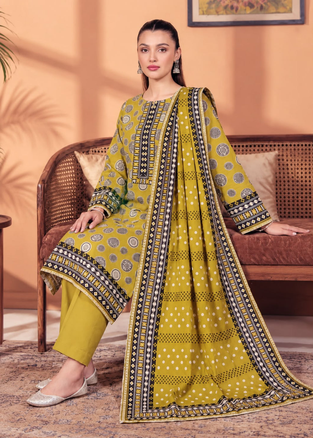 A woman models a three-piece Indian ethnic salwar kameez suit set in a warm, styled indoor setting. The set features a mustard yellow kurta with black and silver geometric patterns, matching yellow trousers, and a coordinating dupatta with a wide black and silver border.