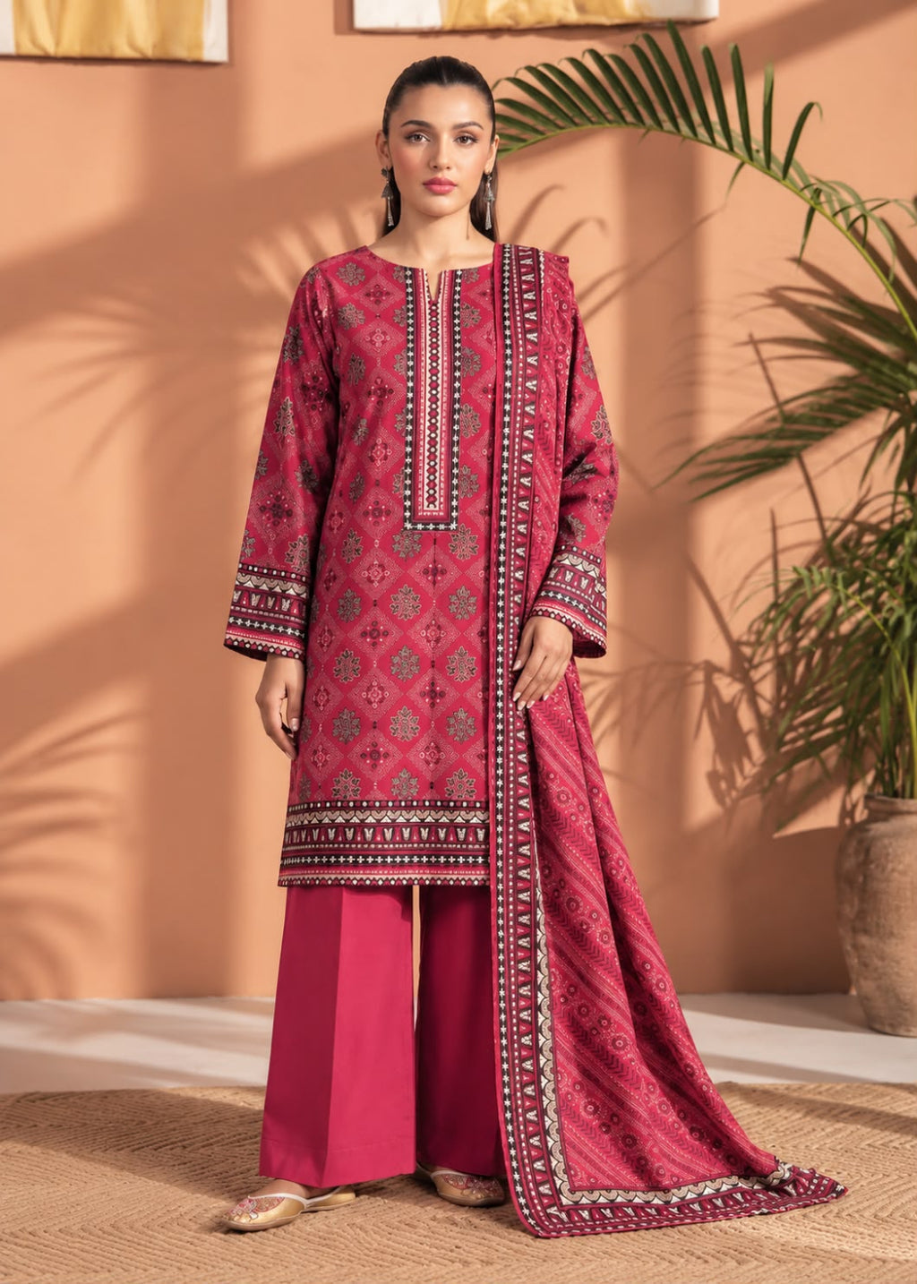 A woman models a three-piece traditional South Asian salwar kameez suit in a warm, earthy indoor setting. The set features a deep magenta-pink kurta with intricate geometric and floral patterns, matching wide-leg trousers, and a coordinating dupatta with a wide decorative border.