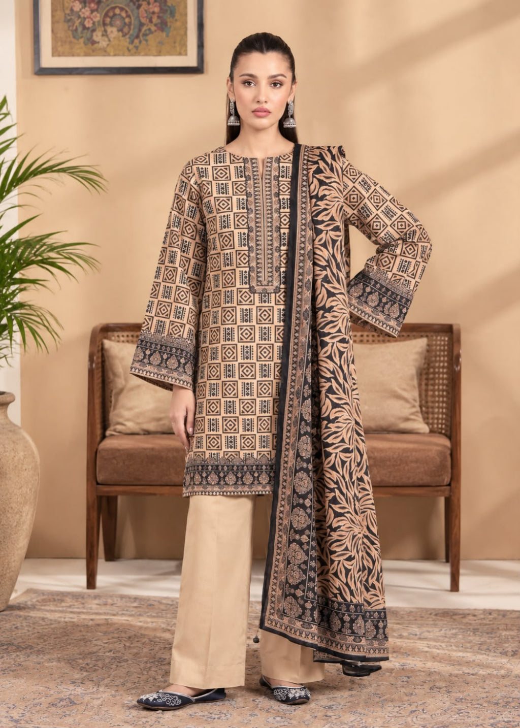 A woman models a traditional three-piece salwar kameez suit in a warm beige tone with intricate black and brown geometric patterns, paired with straight-leg trousers and a matching printed dupatta, set against a cozy indoor background with a wooden chair and potted plant.