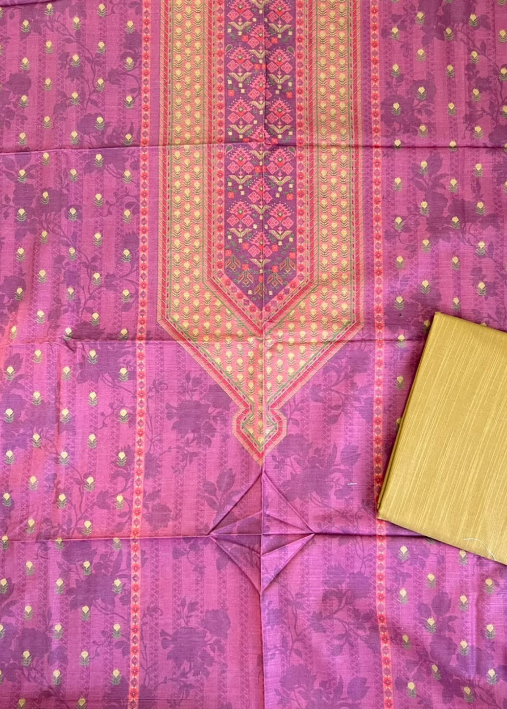A pink and purple patterned fabric laid flat, featuring a central embroidered panel and small gold dot motifs, with a folded matching yellow-gold fabric piece placed in the lower right corner.
