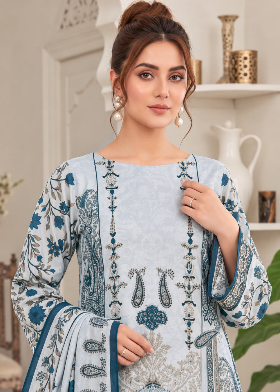 A woman models a light blue and white printed traditional outfit with blue and gray paisley and floral embroidery, featuring a matching dupatta, set against an elegant indoor background.