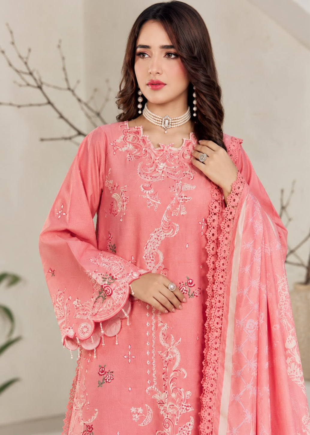A woman models a beige traditional embroidered outfit with brown floral and geometric embroidery, featuring a matching dupatta with a black and white patterned border, set against a softly blurred indoor background.