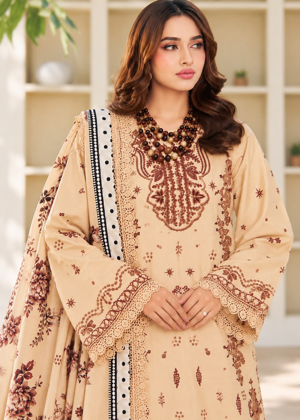 A woman models a beige traditional embroidered outfit with brown floral and geometric embroidery, featuring a matching dupatta with a black and white patterned border, set against a softly blurred indoor background.