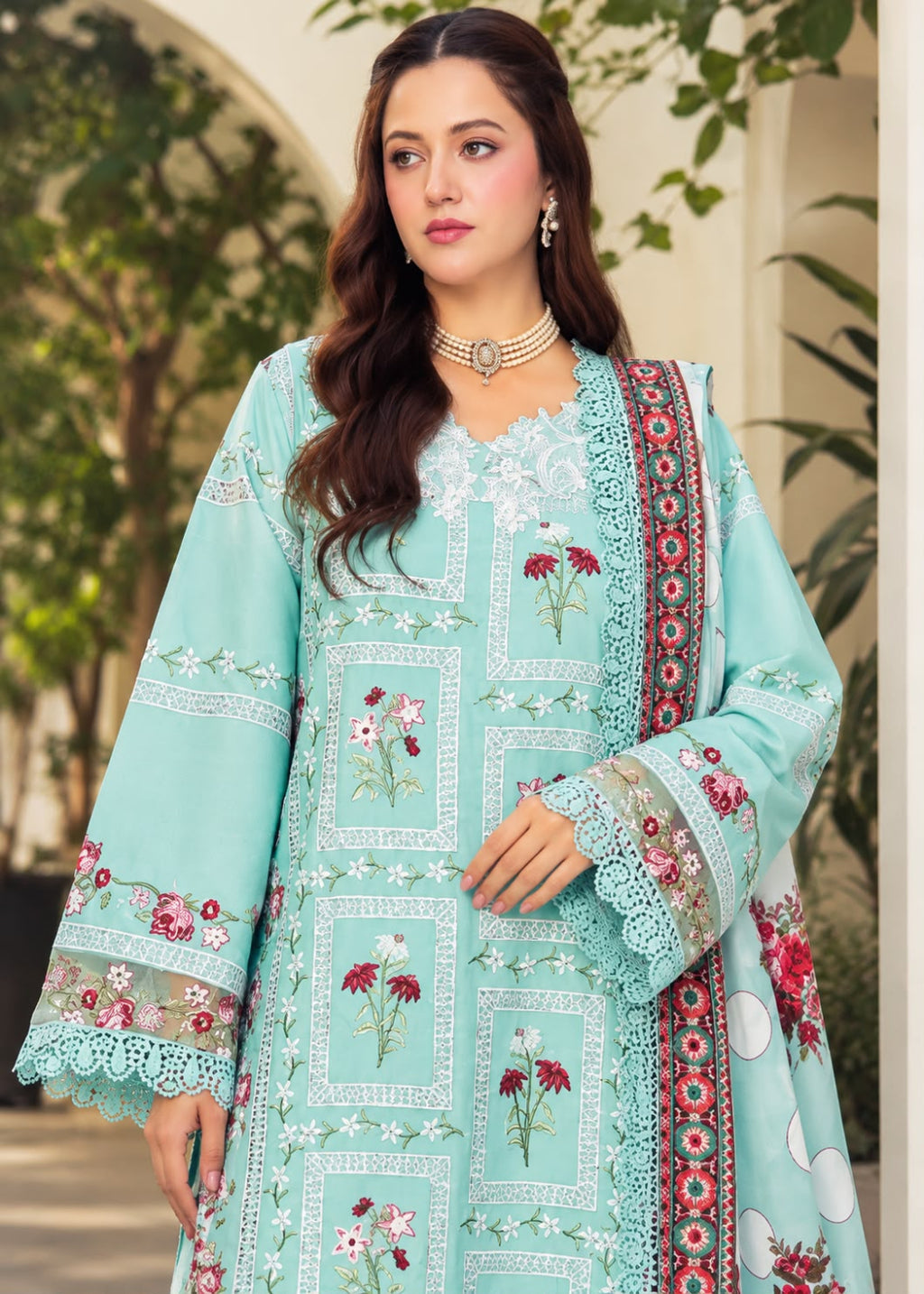 A woman models a beige traditional embroidered outfit with brown floral and geometric embroidery, featuring a matching dupatta with a black and white patterned border, set against a softly blurred indoor background.