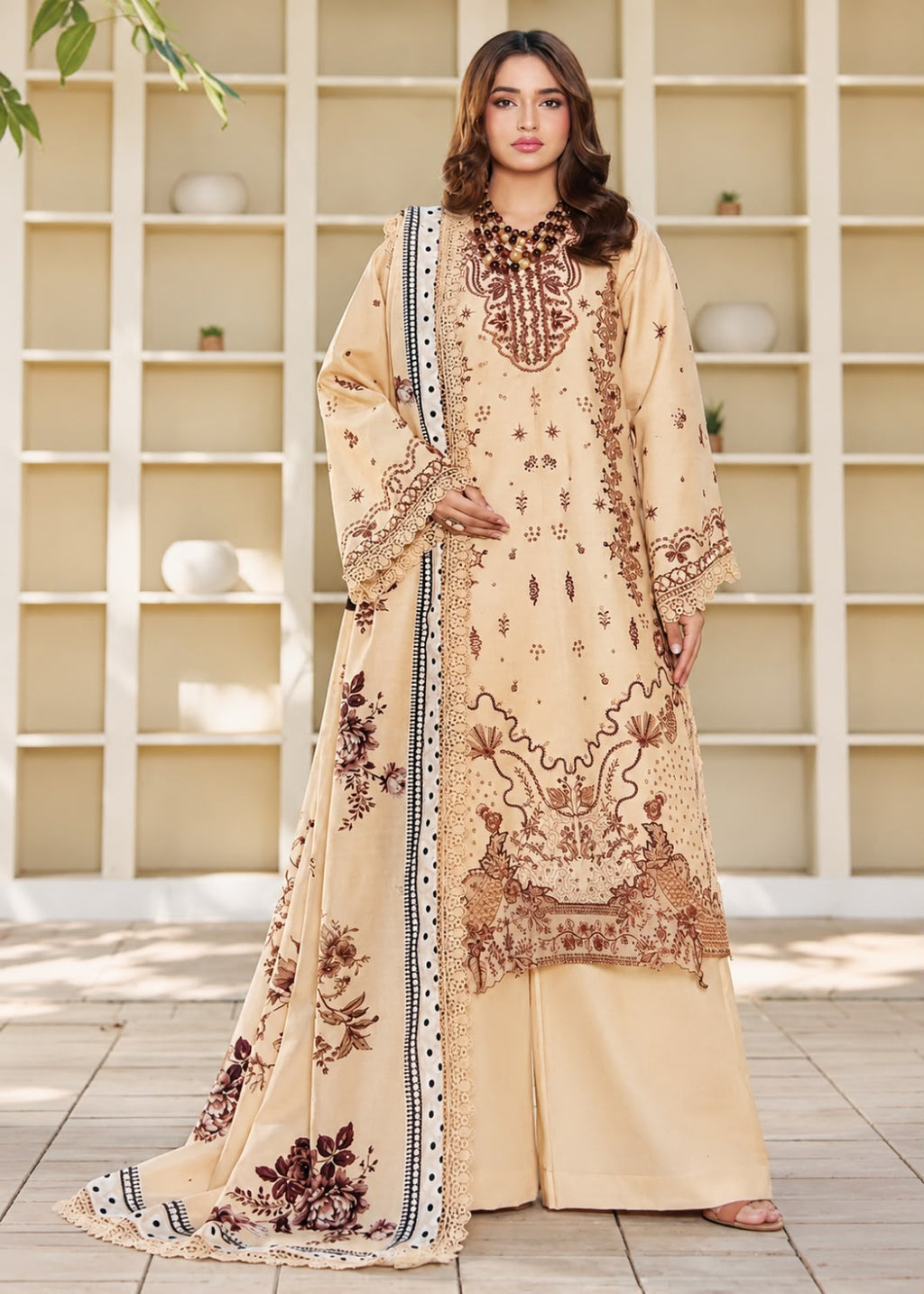 A woman models a traditional three-piece beige ethnic suit with brown floral embroidery and a matching dupatta, set against a beige wooden grid wall background.