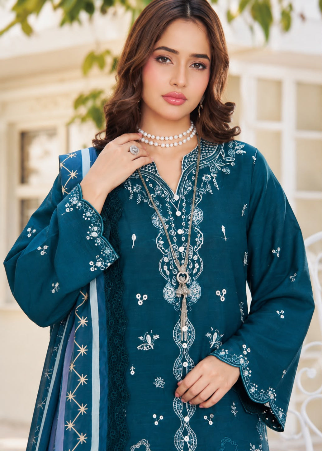 A woman models a beige traditional embroidered outfit with brown floral and geometric embroidery, featuring a matching dupatta with a black and white patterned border, set against a softly blurred indoor background.