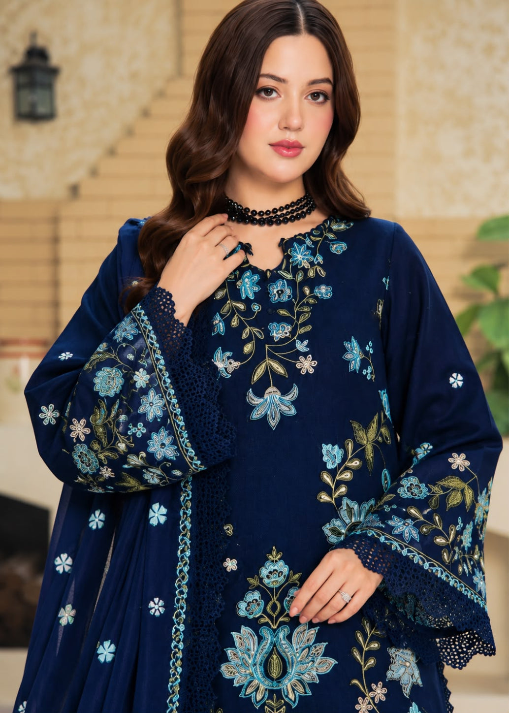A woman models a beige traditional embroidered outfit with brown floral and geometric embroidery, featuring a matching dupatta with a black and white patterned border, set against a softly blurred indoor background.