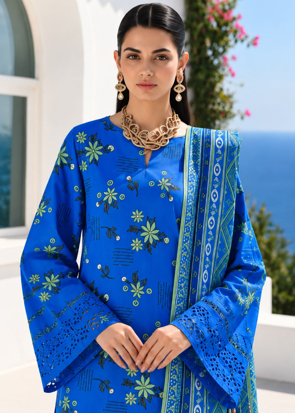 A woman modeling a royal blue printed ethnic kurta set with a matching dupatta, featuring green and yellow floral motifs and intricate embroidery, set against a sunny outdoor background with white walls and blue sea.