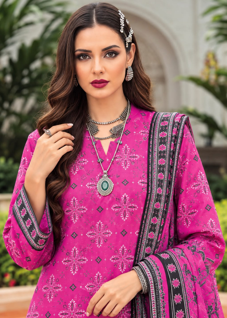 A woman modeling a bright pink traditional outfit with intricate silver and black printed patterns, accessorized with silver jewelry and a patterned dupatta, set against a blurred outdoor background.