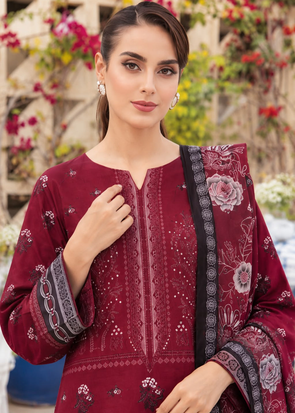A woman wearing a deep maroon embroidered salwar kameez suit with a matching printed dupatta, set against a blurred outdoor background of green foliage and flowers.