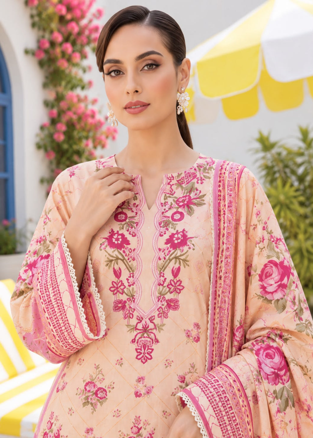 A woman modeling a peach-colored traditional embroidered outfit with pink floral patterns and matching dupatta, set against a bright, sunny outdoor background with yellow umbrellas and green foliage.