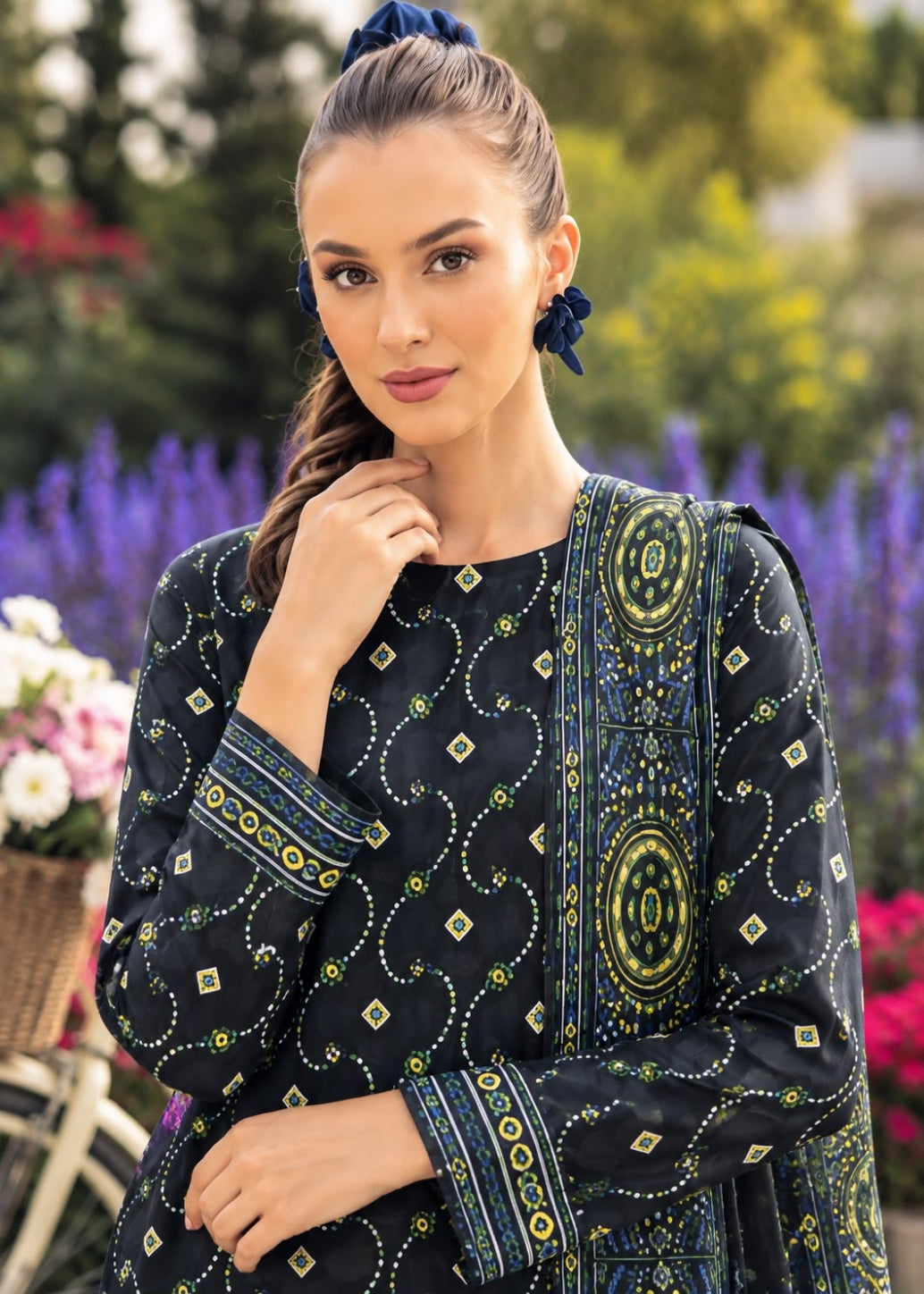 A woman wearing a black embroidered kurta with blue and yellow patterns, posing outdoors in front of a blurred background of purple flowers and green foliage.