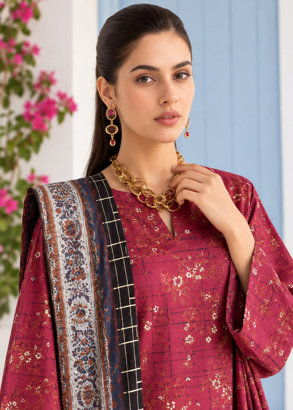A woman modeling a traditional Indian salwar kameez suit in magenta with gold floral prints, featuring a navy blue and gold patterned dupatta, gold jewelry, and a blue paneled background.