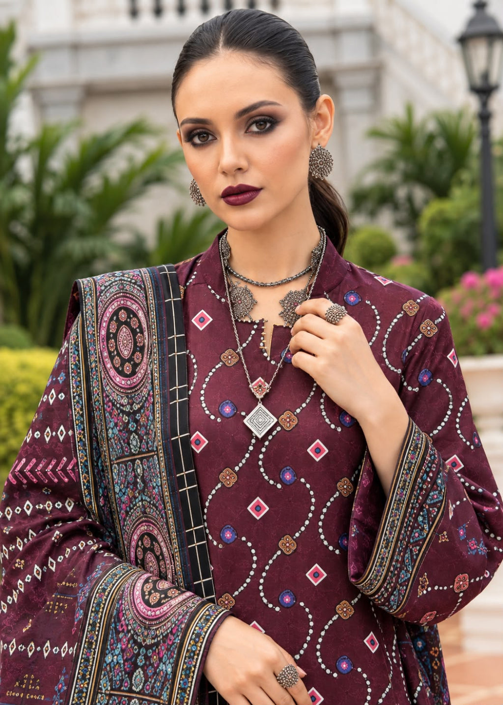 A woman modeling a traditional embroidered kurta in deep maroon with intricate multicolor geometric and floral patterns, paired with matching patterned dupatta and silver jewelry, set against a blurred outdoor garden background.