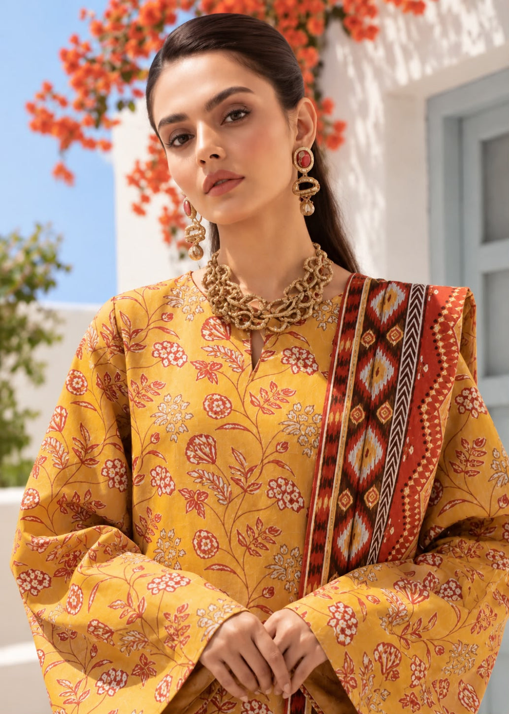 A woman wearing a mustard yellow printed kurta with red and pink floral motifs and a red patterned dupatta, styled with gold jewelry in an outdoor setting with white walls and orange flowers.
