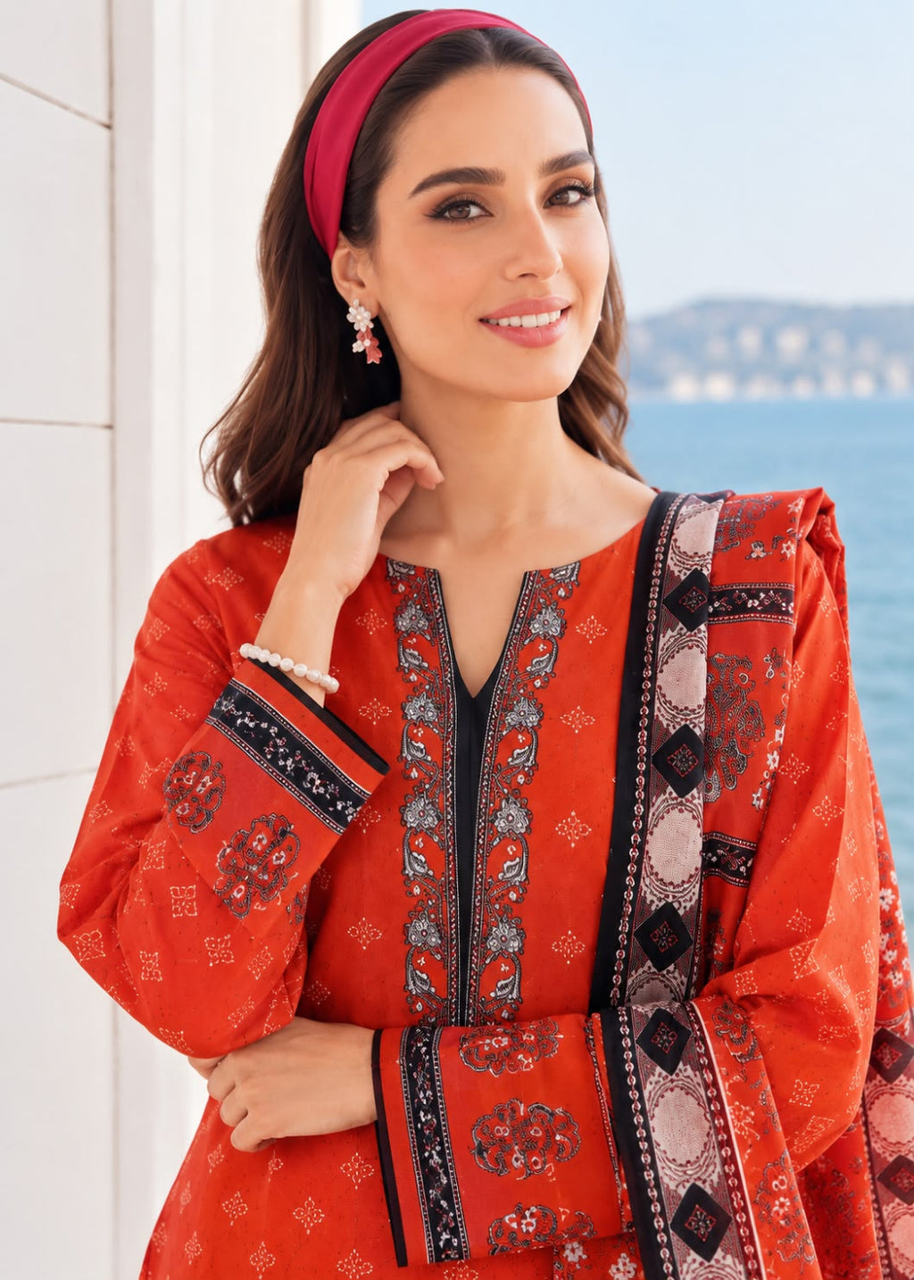 A woman wearing a bright orange-red embroidered kurta with a matching dupatta and pink headband, posed outdoors near a white wall and water.