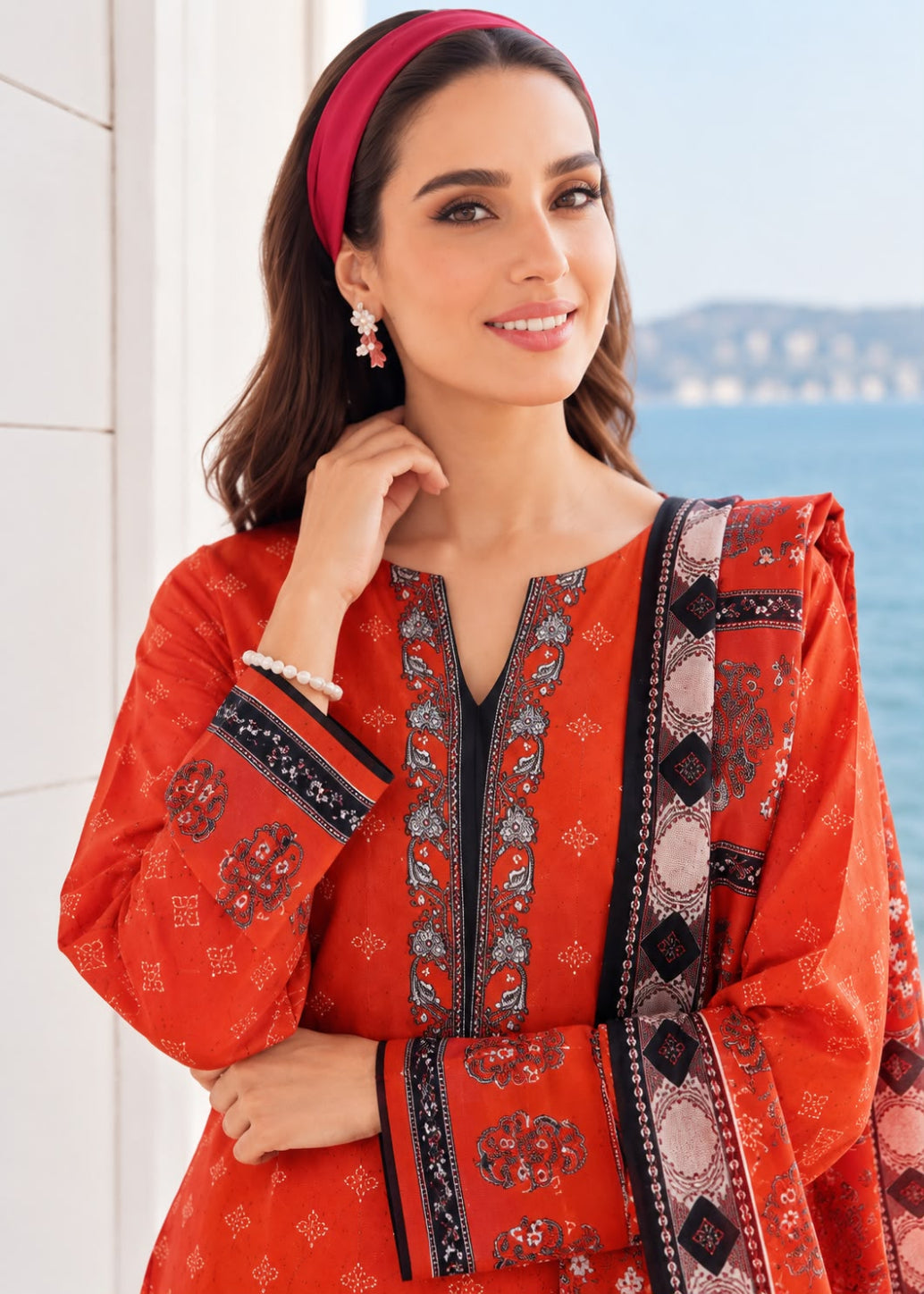 A woman wearing a bright orange-red embroidered kurta with a matching dupatta and pink headband, posed outdoors near a white wall and water.