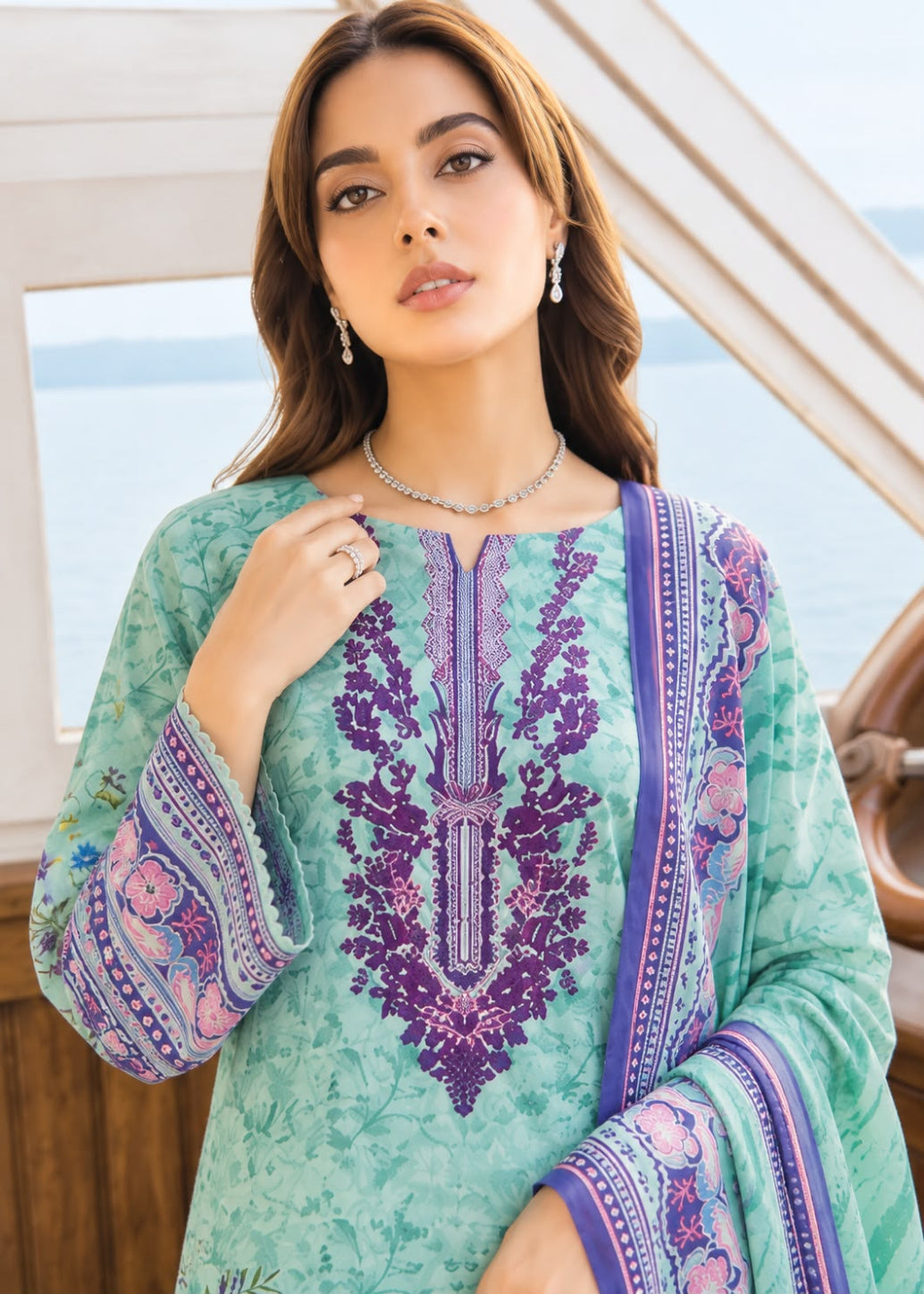 A woman wearing a mint green embroidered kurta with purple and pink floral patterns, paired with a matching dupatta, set against a background of a boat deck and water.