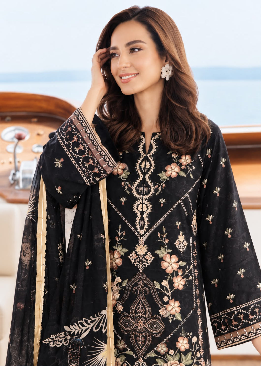 A woman wearing a black embroidered traditional outfit with floral patterns and a matching dupatta, posed on a boat with water and wooden railing visible in the background.