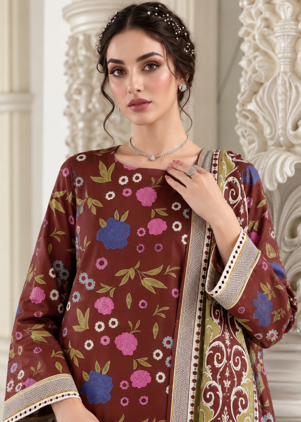 A woman models a brown three-piece traditional outfit with a colorful floral print and ornate patterned borders, set against a classical white stone column background.