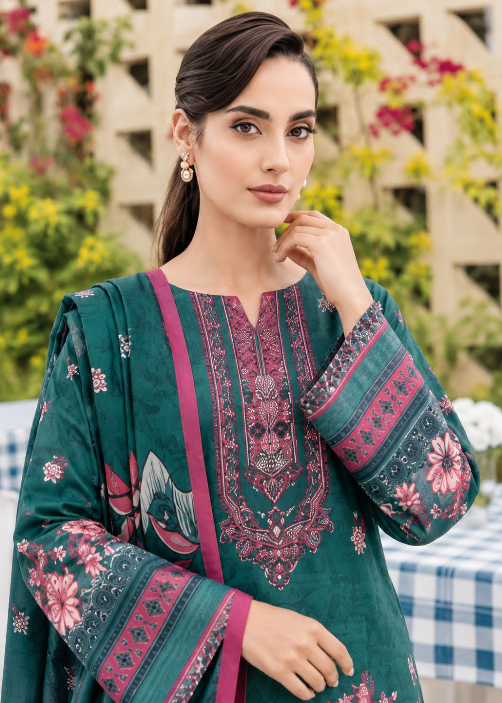 A woman wearing a dark teal green traditional kurta with pink and magenta floral embroidery, set against an outdoor garden background.