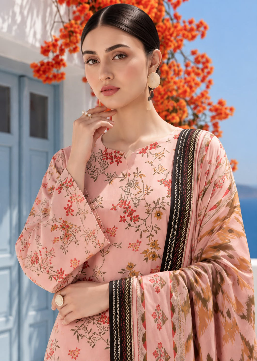 A woman models a light pink floral salwar kameez suit with a matching dupatta featuring black and gold borders, set against a background of a blue door and orange flowers.