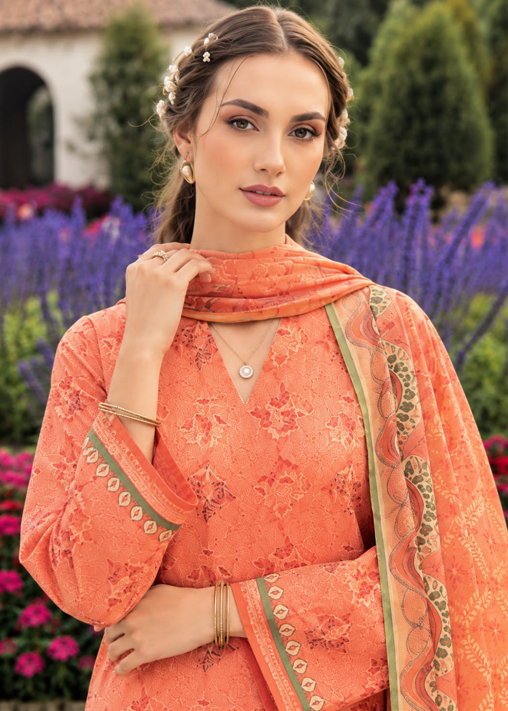 A woman wearing a peach-orange embroidered traditional outfit with a matching dupatta, posed outdoors in front of purple flowers and green foliage.