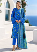 A woman models a royal blue Indian ethnic salwar kameez suit with green floral embroidery and a matching patterned dupatta, set against a white Mediterranean-style building and blue sea background.