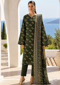A woman models a dark green three-piece ethnic suit with gold and yellow floral prints, matching trousers, and a patterned dupatta, set against a scenic Mediterranean background with white arches and the sea.