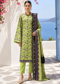 A woman models a three-piece lime green salwar kameez suit with purple and black floral patterns, matching trousers, and a patterned dupatta, set against a Mediterranean background with the sea and blue sky.