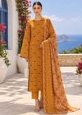 A woman models a mustard yellow ethnic suit set featuring a long kurta with intricate embroidery, matching trousers, and a coordinating printed dupatta, set against a Mediterranean-style outdoor background with the sea in the distance.