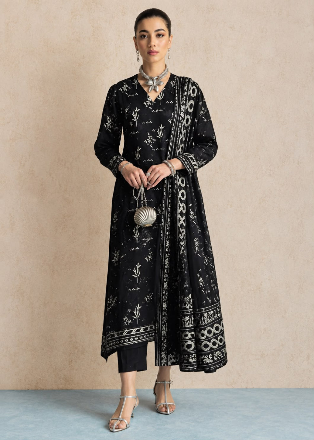 A woman models a black long kurta with white floral and geometric block prints, paired with black pants, silver sandals, and a small silver purse, standing against a textured beige wall.