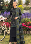 A woman stands outdoors beside a vintage bicycle in a garden, wearing a black traditional outfit with intricate gold and green embroidered patterns and a matching draped scarf.